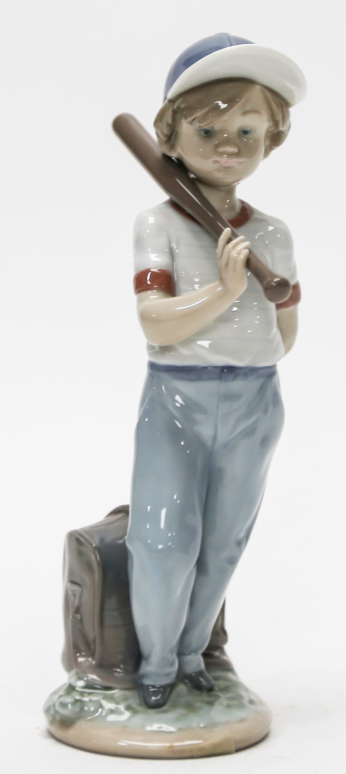 Lladró Young Baseball Player Figurine: Glossy Lladró porcelain figurine depicting a young baseball player wearing a blue cap and uniform, carrying a bat over his shoulder with a baseball held behind his back. A detailed equipment bag rest