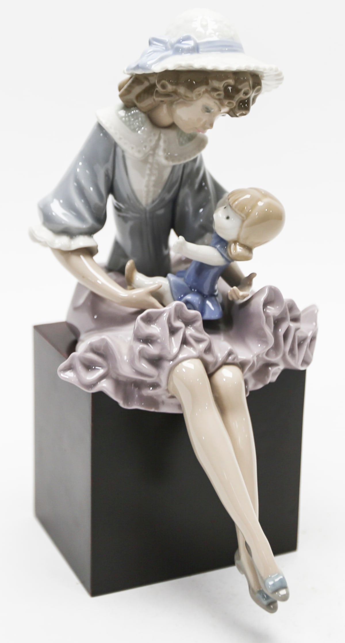 Lladro Mother and Child Porcelain Figurine on Wood Base: A Lladro glazed porcelain figurine depicting a seated mother wearing a blue blouse, lavender ruffled skirt, and wide-brimmed hat while holding a young child on her lap. Rendered in the makers