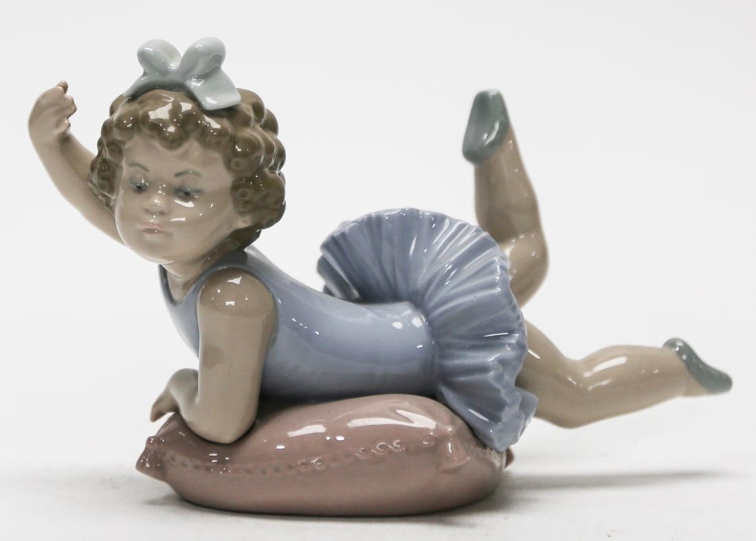 Lladro Reclining Ballerina on Cushion Porcelain Figurine: A retired Lladro glazed porcelain figurine depicting a young ballerina reclining playfully on a pink cushion, wearing a pale blue leotard and ruffled tutu with a matching bow in her curled hair. Handm