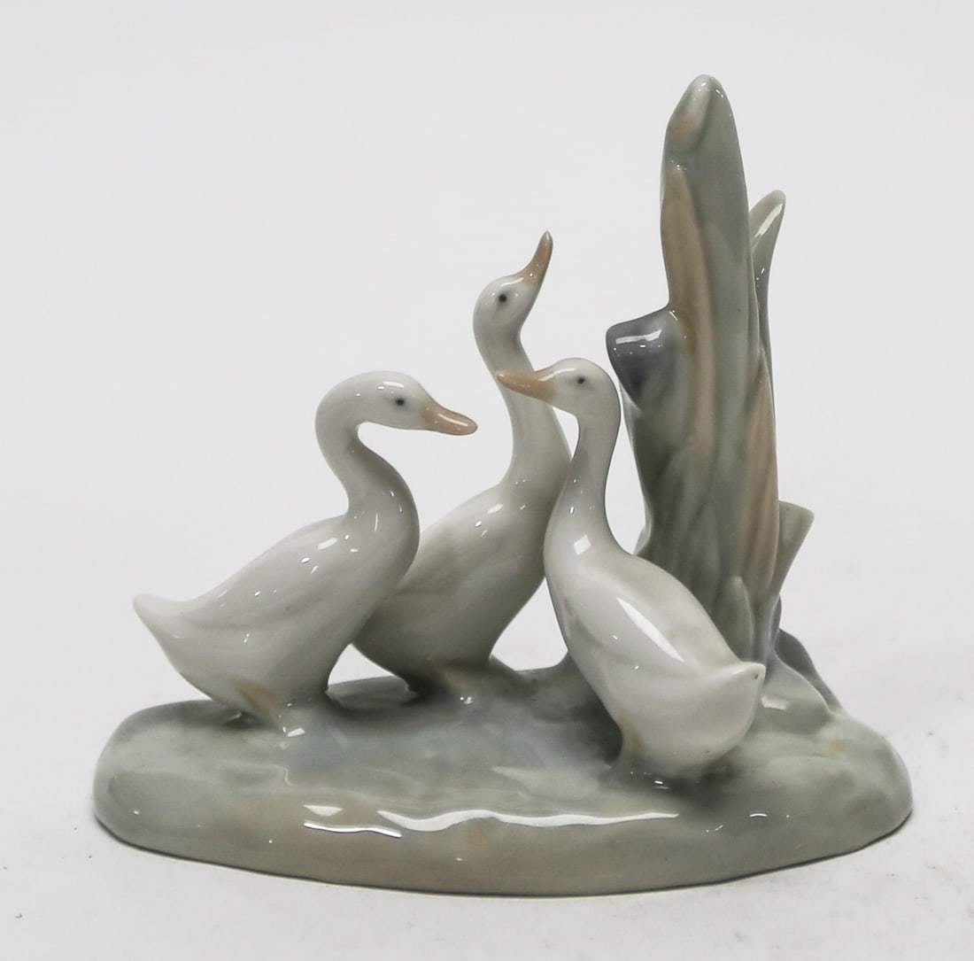 Spanish Porcelain Three Geese Figurine with Reeds: Glossy porcelain figurine featuring three geese gathered beside tall reeds in soft gray and beige tones. Marked handmade in Spain on the base. Measures approximately 5 x 5 inches. Pre-owned with light