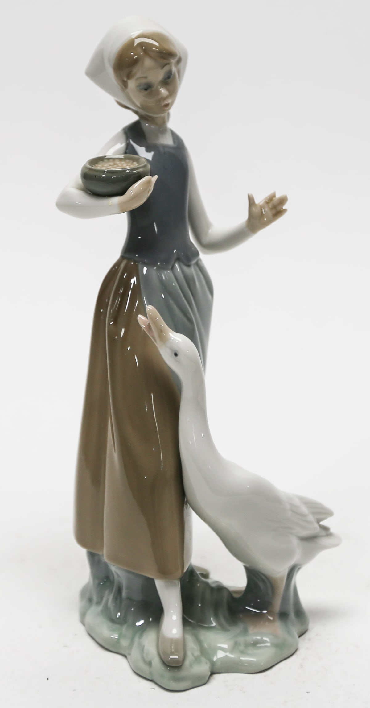 Lladro Girl Feeding Goose Porcelain Figurine: Glossy porcelain figure depicting a young woman holding a bowl while feeding a goose. Finished in muted brown, gray, white, and pale green tones. Lladro blue maker mark on underside. Measures approxim