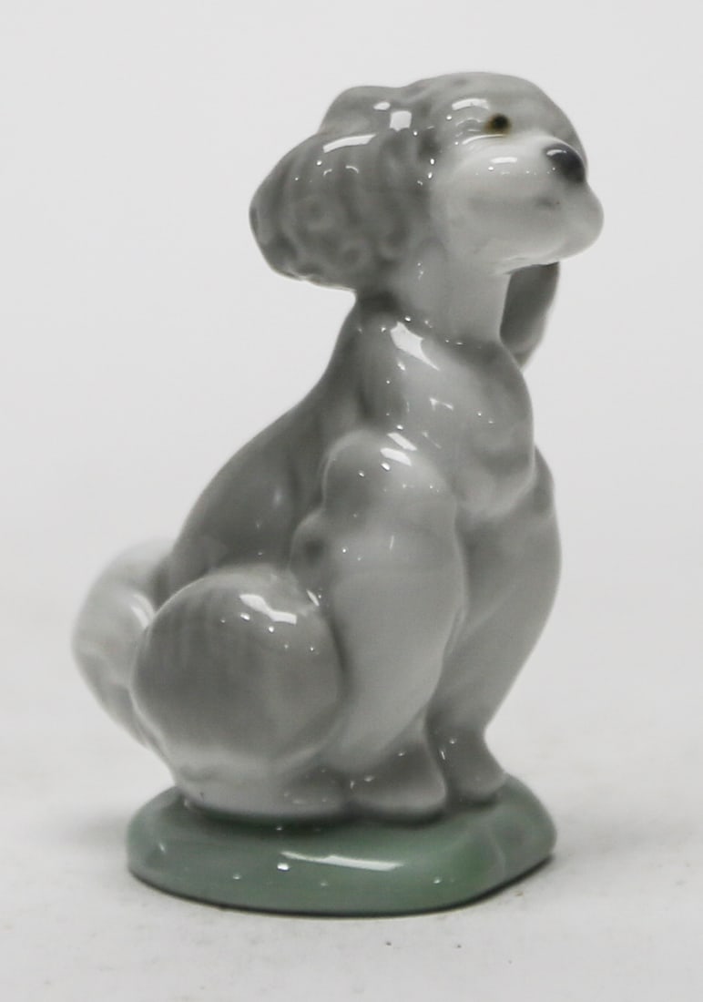 Lladro Society 2000 Seated Dog Porcelain Figurine: Small glazed porcelain figure depicting a seated gray dog on a pale green oval base. Blue Lladro Society 2000 mark underneath. Measures approximately 3 x 2 inches. Good condition with light surface an