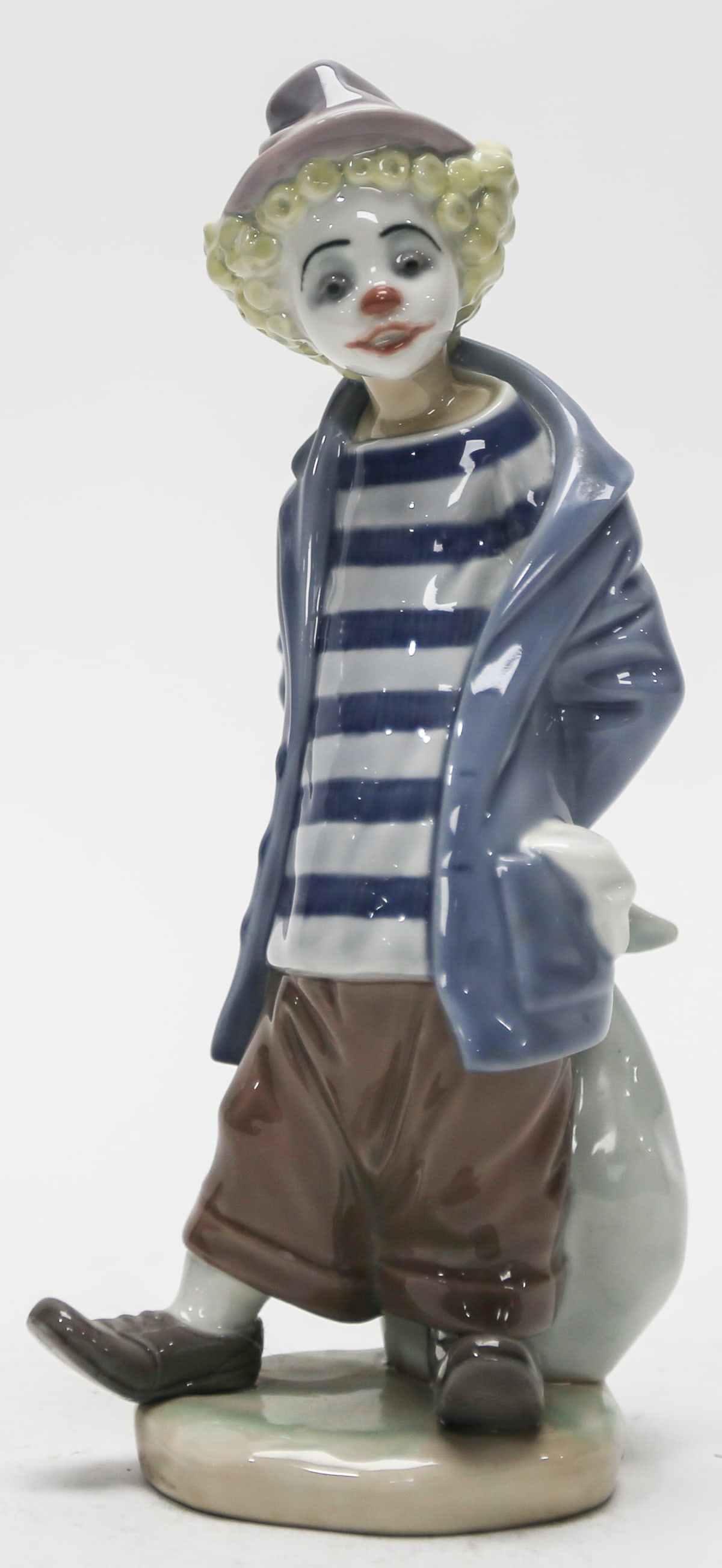 Lladro Little Traveler Clown Porcelain Figurine No. 7602: A retired Lladro glazed porcelain figurine titled Little Traveler, model No. 7602, depicting a contemplative clown walking with an oversized travel sack held behind his back. The figure wears a lavend