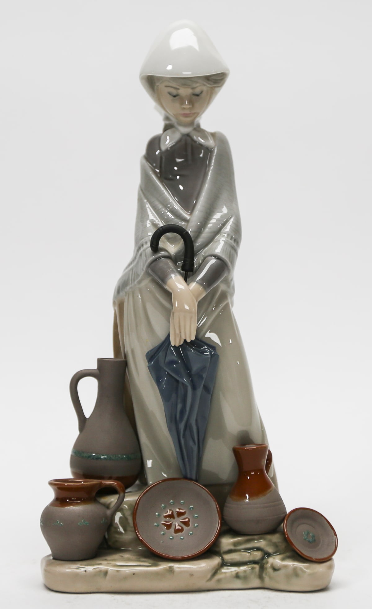 Lladro The Ceramic Seller No. 5080 Porcelain Figurine: Large Spanish glazed porcelain figurine depicting a young ceramic seller wearing a white headscarf, gray fringed shawl, dark bodice, and long neutral-toned skirt. She holds a closed blue umbrella whil