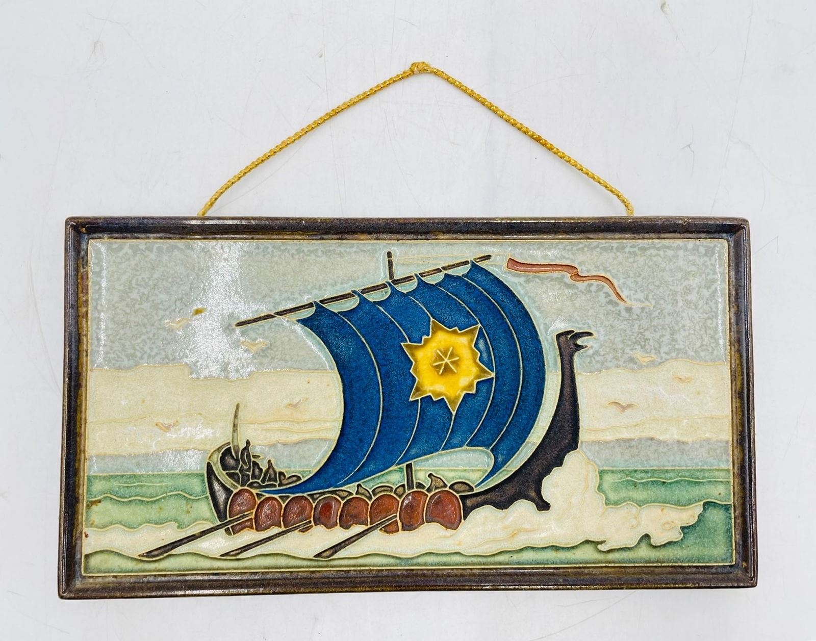 Hand-Glazed Dutch Delft Ceramic Tile Plaque Depicting Viking Longship – Marked: A striking mid-century Dutch Delft ceramic wall plaque depicting a stylized Viking longship sailing across ocean waves. The composition features a bold cobalt blue sail adorned with a yellow starburst