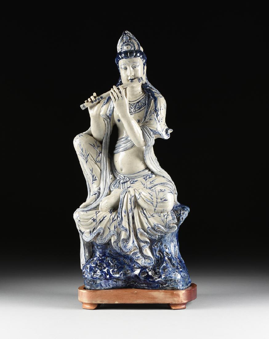 A MING DYNASTY (1368-1644) STYLE BLUE AND WHITE: A MING DYNASTY (1368-1644) STYLE BLUE AND WHITE STONEWARE SCULPTURE OF THE GUANYIN PLAYING FLUTE, 19TH/20TH CENTURY, the goddess of compassion sits on a blue rock with scrolling head dress over a curl