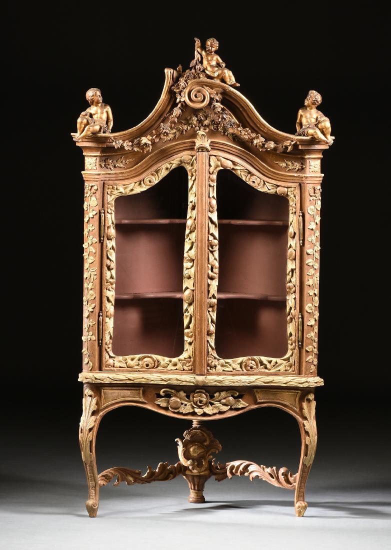 A LARGE ITALIAN ROCOCO STYLE PARCEL GILT AND PAINTED: A LARGE ITALIAN ROCOCO STYLE PARCEL GILT AND PAINTED CARVED WOOD CORNER VITRINE, 18TH AND 19TH CENTURY, the scrolling broken arched cornice surmounted by three seated putti holding aloft nicely detail