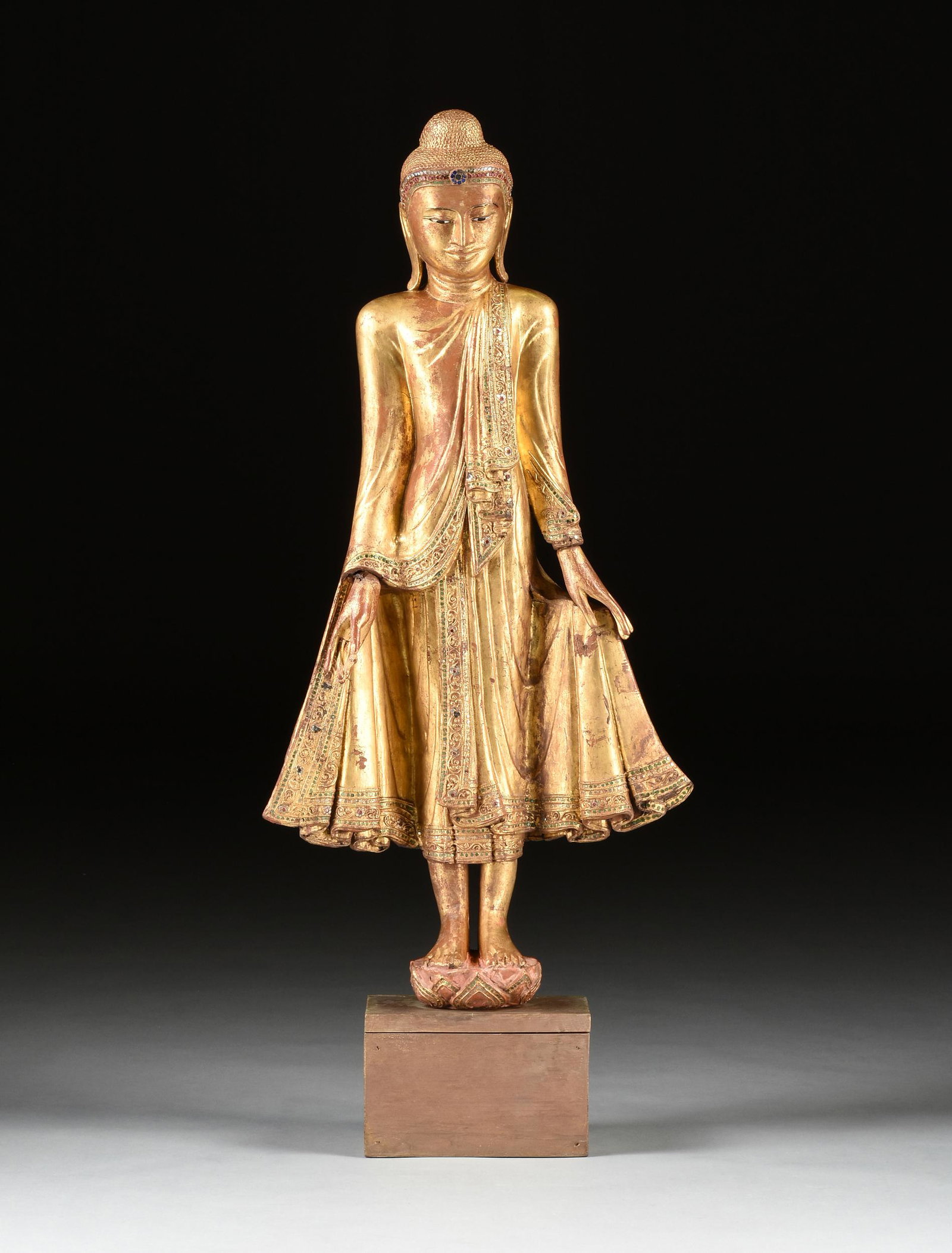 A BURMESE FAUX JEWELED GILTWOOD FIGURE OF BUDDHA, LATE: A BURMESE FAUX JEWELED GILTWOOD FIGURE OF BUDDHA, LATE 20TH CENTURY, carved standing in samabhanga on a short stylized lotus base and wearing an ushnisha on a tightly curled hairstyle before a mica an
