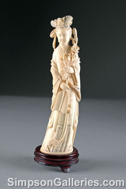 A LATE QING DYNASTY CARVED IVORY FIGURE OF QUAN YI: A LATE QING DYNASTY CARVED IVORY FIGURE OF QUAN YIN, modeled with right hand holding a bouquet of flowers and her left hand holding a fan, with elaborate avial headdress. On custom hardwood stand. Hei