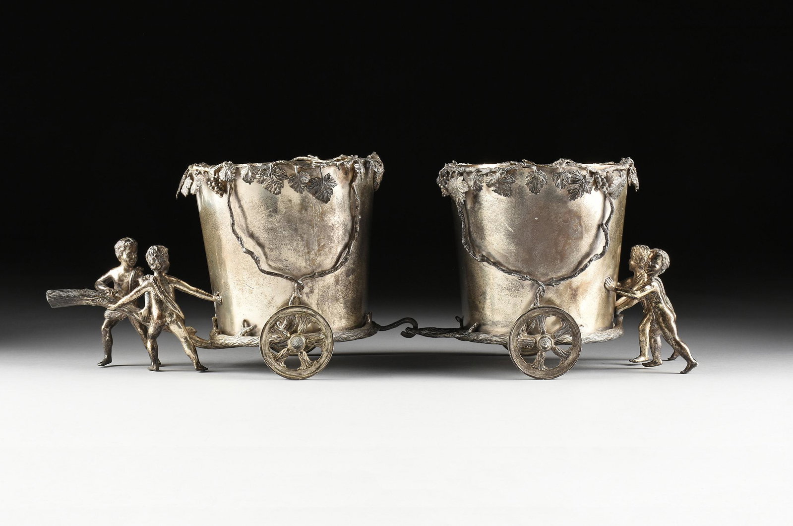 A MAITLAND-SMITH SILVERPLATED TABLETOP WINE BUCKETS: A MAITLAND-SMITH SILVERPLATED TABLETOP WINE BUCKETS TROLLEY, MODERN, composed of two wine cooler buckets and each mounted within a grapevine and clusters frame on wheels, joined by grapevine form hook