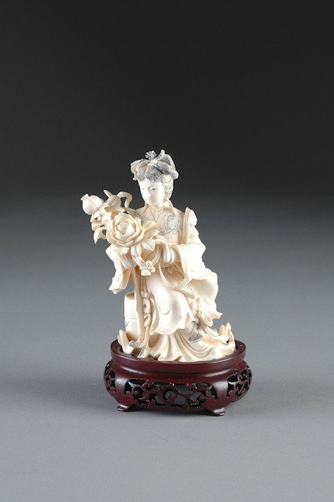 A CHINESE CARVED IVORY FIGURE OF A YOUNG MAIDEN in : A CHINESE CARVED IVORY FIGURE OF A YOUNG MAIDEN in long flowing robes holding a bouquet of flowers and seated on a garden stool. On carved hardwood stand. Height: 6"