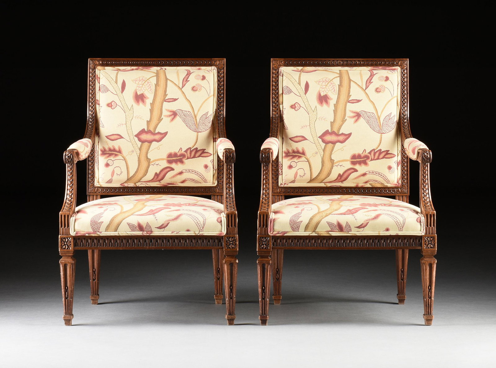 A PAIR OF LOUIS XVI STYLE CARVED AND UPHOLSTERED WALNUT FAUTEUILS, 20TH CENTURY,: A PAIR OF LOUIS XVI STYLE CARVED AND UPHOLSTERED WALNUT FAUTEUILS, 20TH CENTURY, each with a rectangular "monnaie" carved back frame continuing to padded arms with scrolling handrests and continuing t
