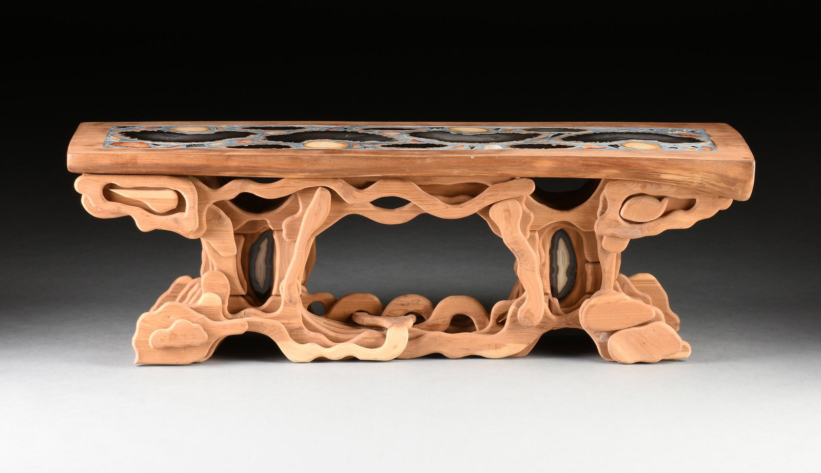A PETRIFIED PALM INLAID AND CARVED CEDAR COFFEE TABLE / BENCH, 1970s,: A PETRIFIED PALM INLAID AND CARVED CEDAR COFFEE TABLE / BENCH, 1970s, the long rectangular top centering a blue composite panel inlaid with horizontally cut samples of various colorful petrified palm 