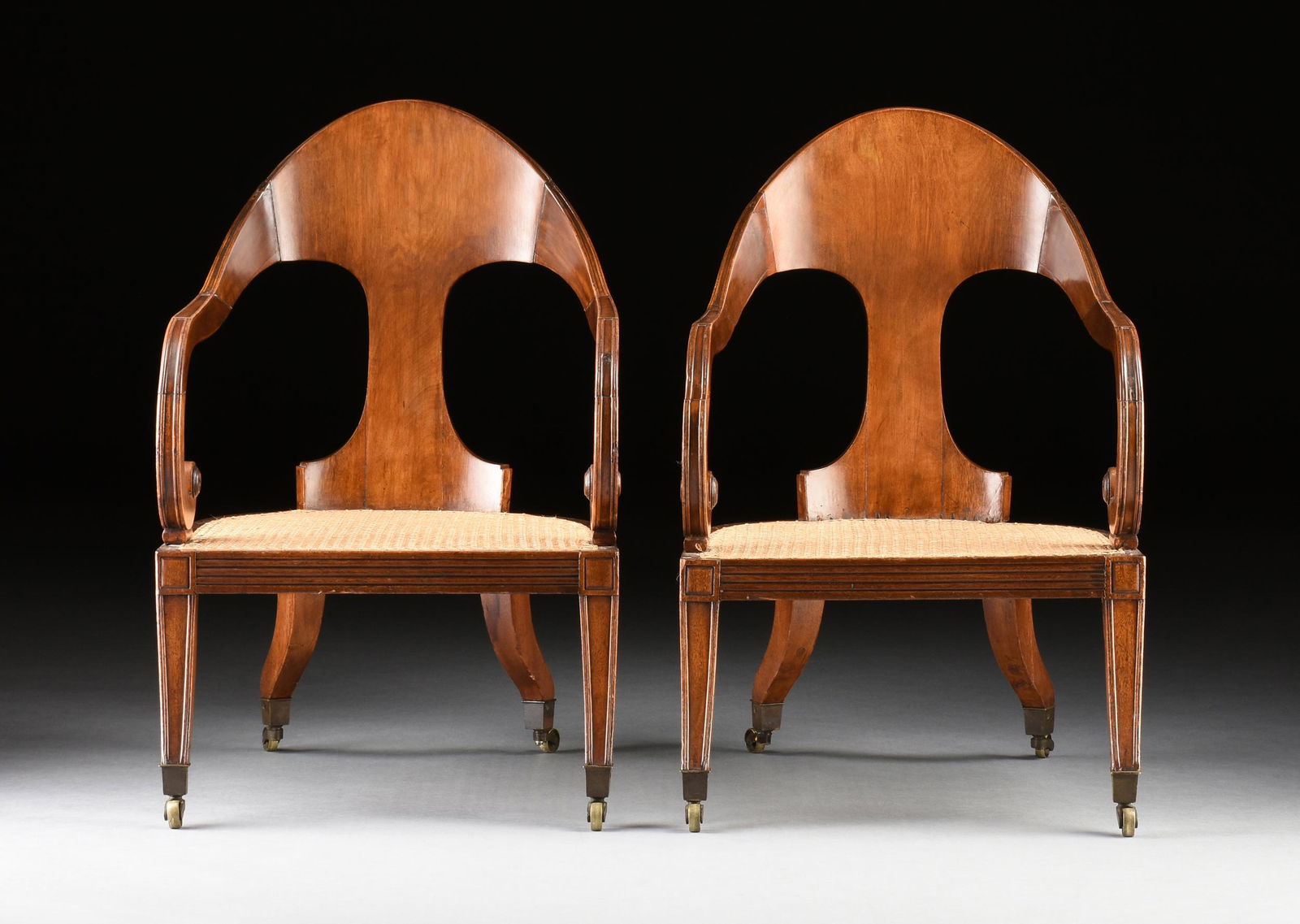 A PAIR OF REGENCY CARVED MAHOGANY AND CANED GONDOLA ARMCHAIRS, EARLY 19TH CENTURY,: A PAIR OF REGENCY CARVED MAHOGANY AND CANED GONDOLA ARMCHAIRS, EARLY 19TH CENTURY, each with an arched and channeled crestrail above a broad back tapering to a narrow vertical splat, flanked by scroll