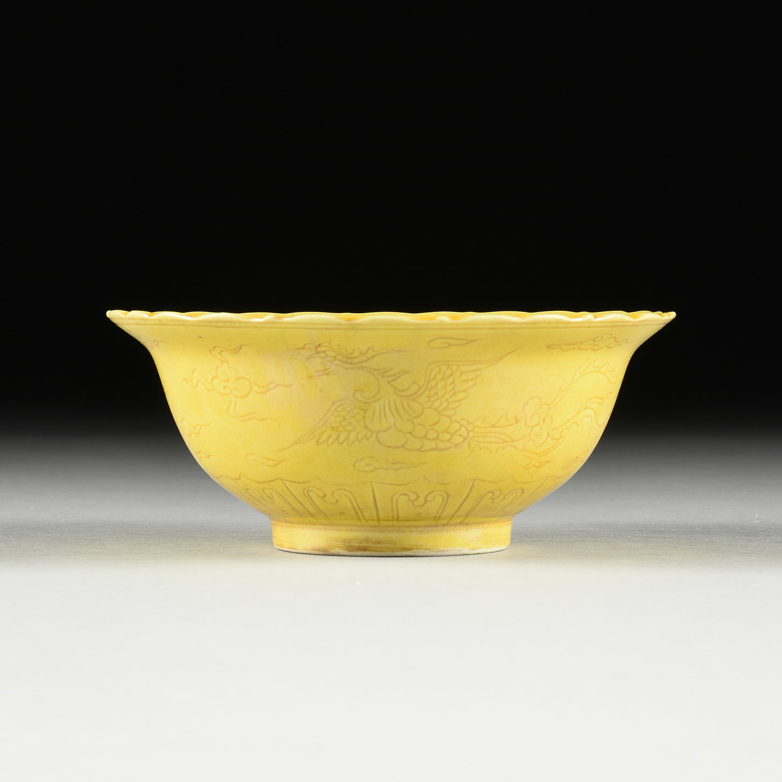 A MING DYNASTY STYLE EGG YOLK YELLOW GLAZE "PHOENIX" INCISED BOWL, 20TH CENTURY,: A MING DYNASTY STYLE EGG YOLK YELLOW GLAZE "PHOENIX" INCISED BOWL, 20TH CENTURY, the shaped floriform edge over a bowl with central encircled mystical cloud, the body exterior is carved with celestial
