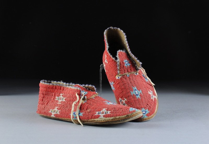 A PAIR OF AMERICAN INDIAN CHEYENNE FULLY BEADED HI: A PAIR OF AMERICAN INDIAN CHEYENNE FULLY BEADED HIDE MOCCASINS, CIRCA 1900, sinew-sewn in red with stylized designs blue edging and beaded tongue, hide ties. Width: 10 1/2"