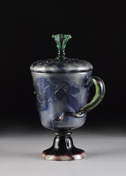 A FINE GALLÉ SIGNED, APPLIED, AND ENAMELED COVERED: A FINE GALLÉ SIGNED, APPLIED, AND ENAMELED COVERED GLASS JAR, the footed vessel in lavender, blue and green glass fire polished with meandering vinery. With conforming cover and applied “C” scrol