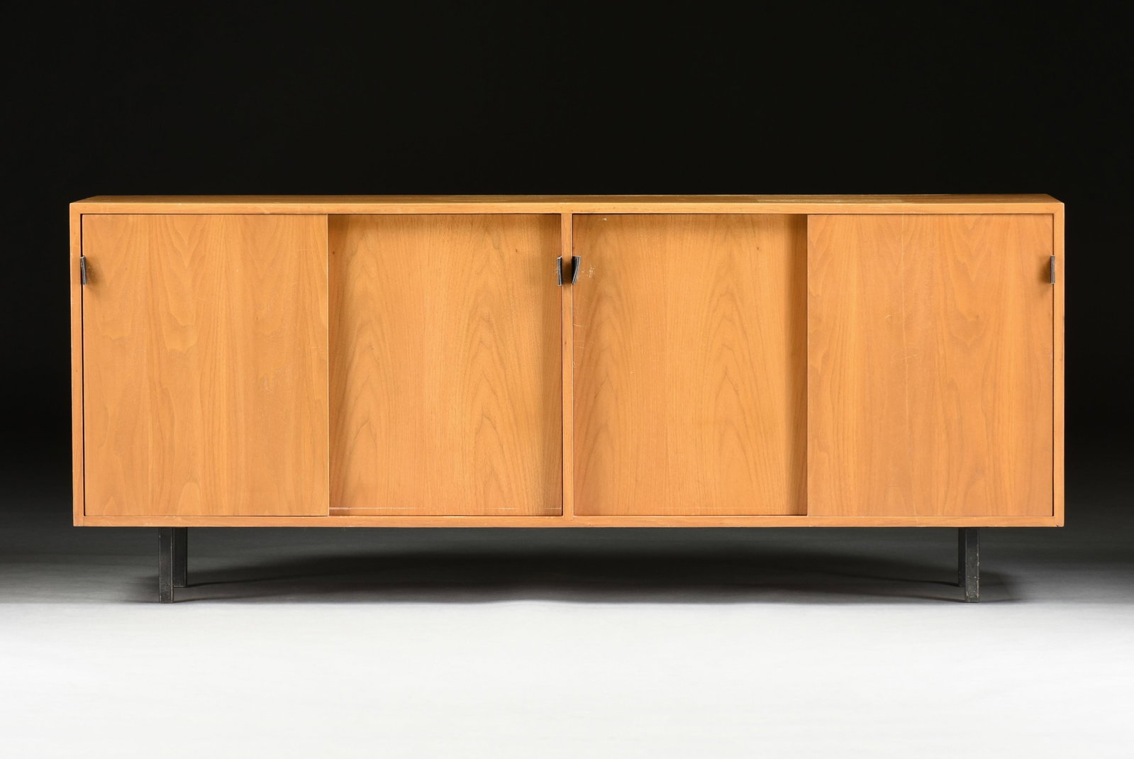 A MID-CENTURY MODERN SLIDING DOOR CREDENZA, 20TH CENTURY: A MID-CENTURY MODERN SLIDING DOOR CREDENZA, 20TH CENTURY, the oak veneer wooden body with four sliding panel doors, each with a leather pull, supported on four metal legs, in the style of Florence Kno