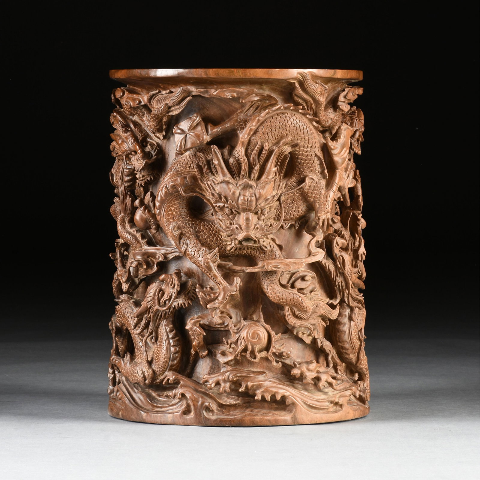 A WOODEN UMBRELLA STAND, CHINESE, 20TH CENTURY, a hardwood umbrella stand hand-carved from a single: A WOODEN UMBRELLA STAND, CHINESE, 20TH CENTURY, a hardwood umbrella stand hand-carved from a single piece of wood and decorated with swirling, writhing dragons. Height: 20 3/4"
