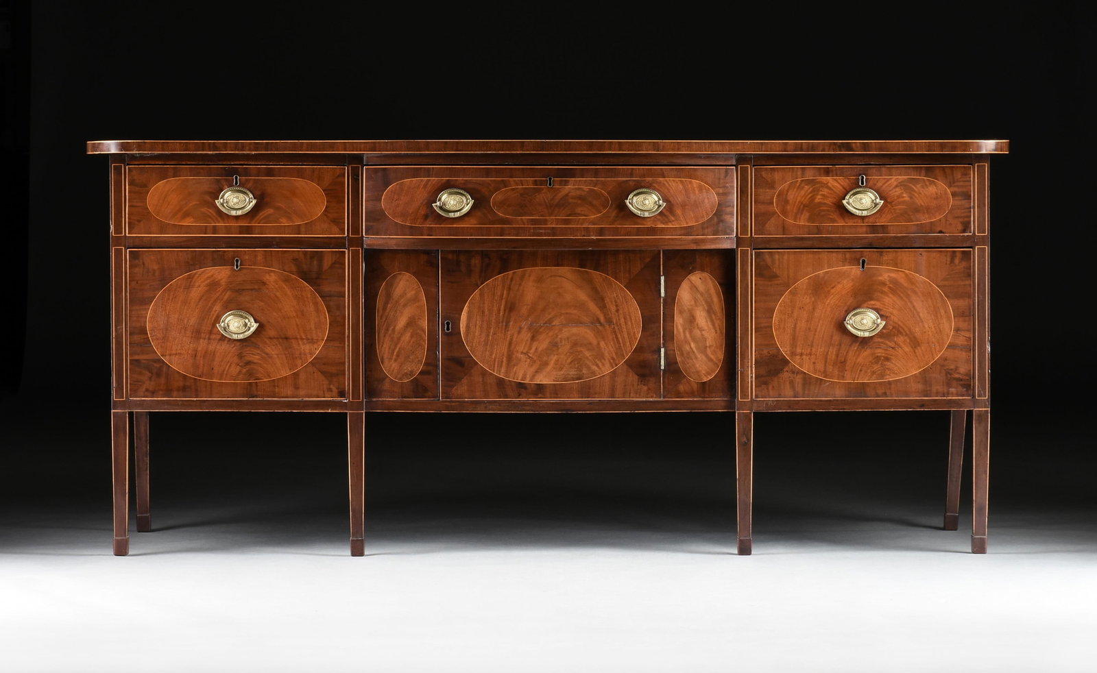 A HEPPLEWHITE STYLE MAHOGANY SIDEBOARD, ENGLISH, CIRCA 1800,: A HEPPLEWHITE STYLE MAHOGANY SIDEBOARD, ENGLISH, CIRCA 1800, a break bow mahogany sideboard with decorative inlay, five drawers and a cabinet with brass hardware, all supported on tapered legs. The lo