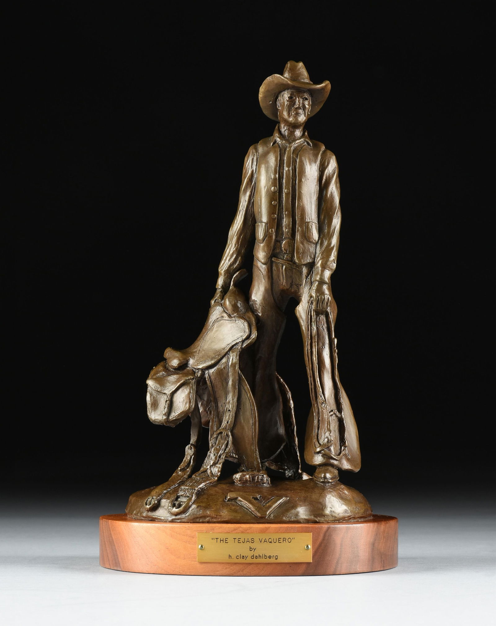 CLAY DAHLBERG (American/Texas b. 1946) "The Tejas Vaquero," 1972,: CLAY DAHLBERG (American/Texas b. 1946) "The Tejas Vaquero," 1972, a patinated bronze sculpture of a man holding a saddle mounted on a wooden base, signed, dated, and numbered, "H. CLAY DAHLBERG ©