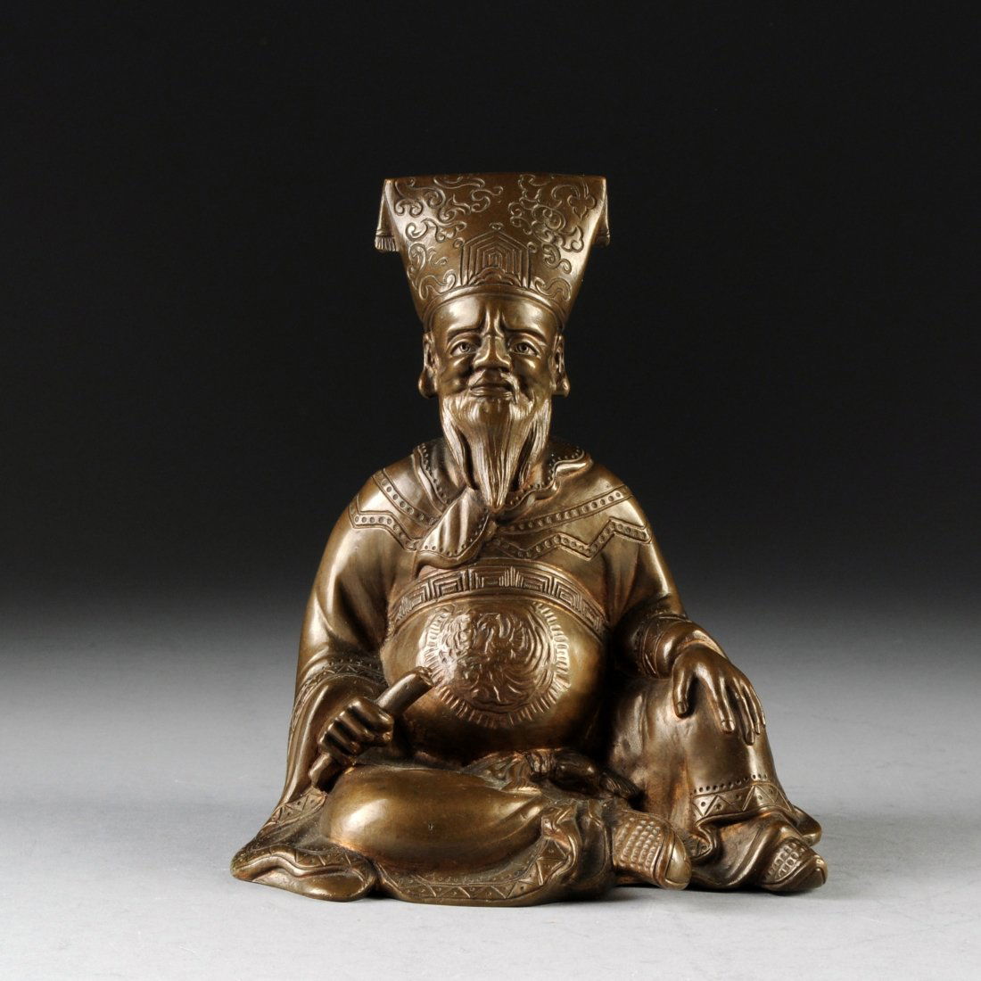 A CHINESE BRONZE SEATED SCHOLAR, LATE 19TH/EARLY 20TH C: A CHINESE BRONZE SEATED SCHOLAR, LATE 19TH/EARLY 20TH CENTURY, the figure holding a scroll in one hand with a tall hat folding over the top, having a long beard and weathered face. Height: 6 3/4" Widt