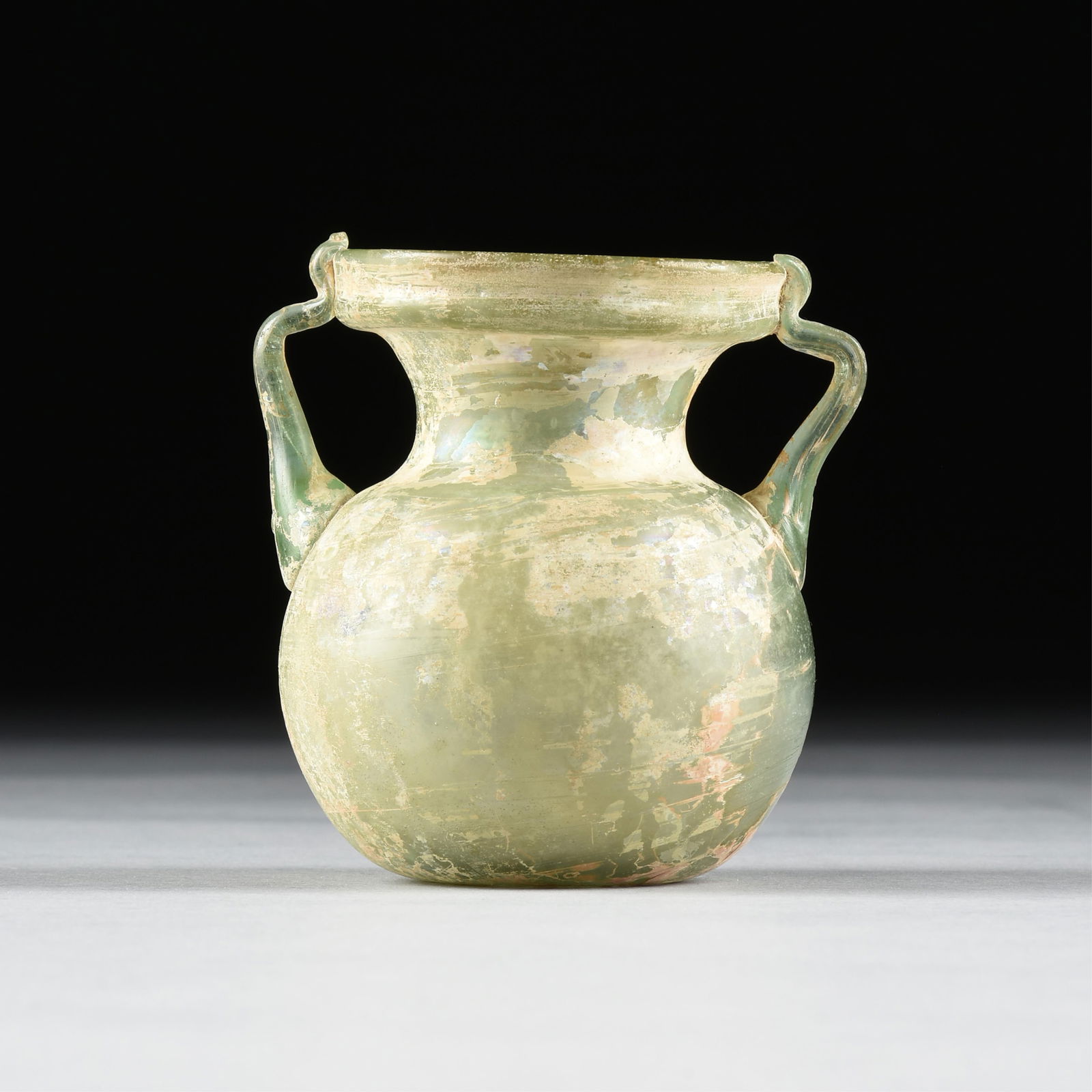 A ROMAN IMPERIAL TWIN-HANDLED GLASS VESSEL, 1ST - 5TH CENTURY,: A ROMAN IMPERIAL TWIN-HANDLED GLASS VESSEL, 1ST - 5TH CENTURY,an iridescent green glass vessel with two handles.4 x 4 x 3 1/2 in. (10.2 x 10.2 x 8.9 cm.) Provenance: Collection of the Norbert Choucrou