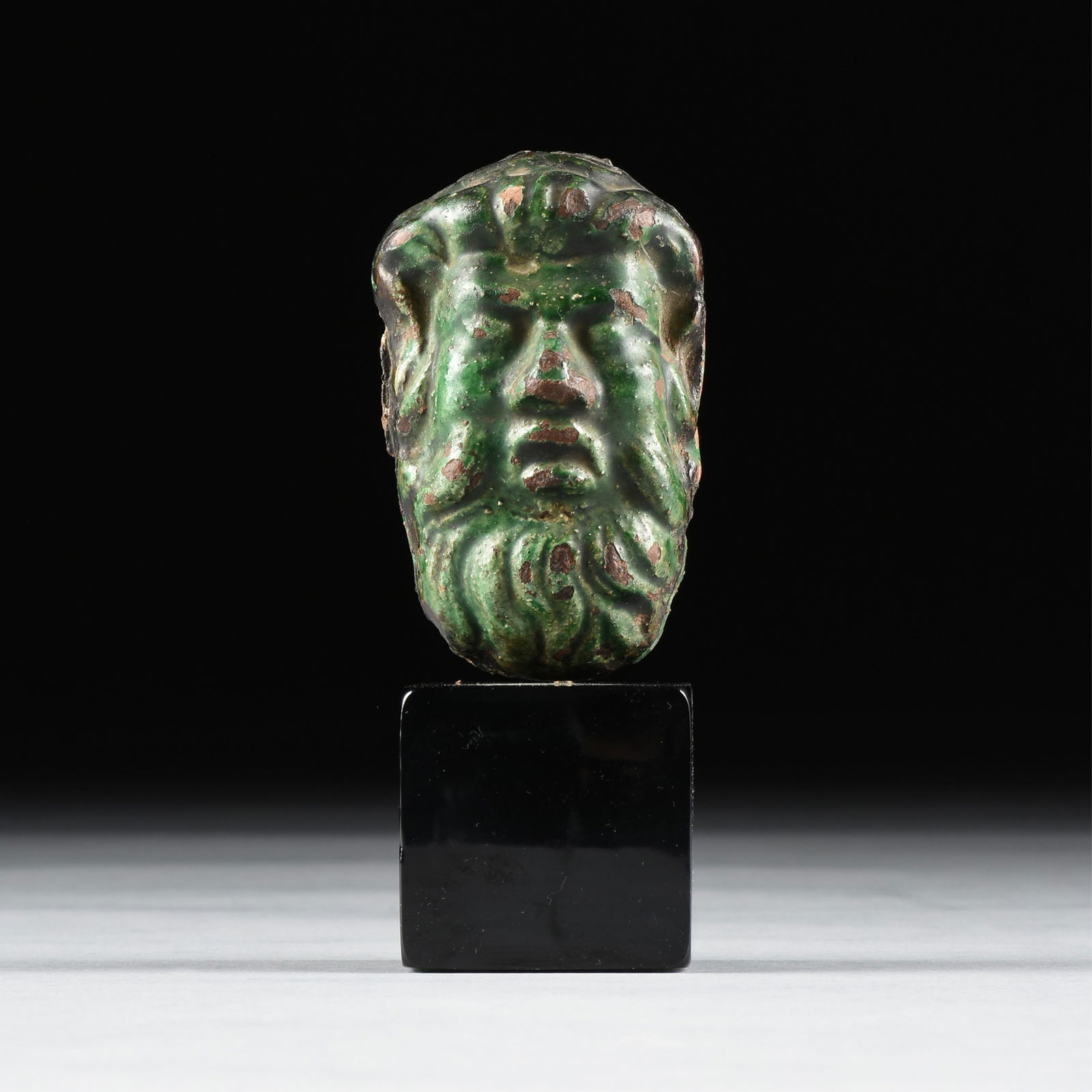 A ROMAN IMPERIAL HEAD OF A GOD, 1ST - 2ND CENTURY CE,: A ROMAN IMPERIAL HEAD OF A GOD, 1ST - 2ND CENTURY CE,a glazed earthenware sculpted head, possibly broken off of a vessel.4 3/4 x 2 3/8 x 1 5/8 in. (12.1 x 6 x 4.1 cm.), With stand: 5 3/4 x 2 3/8 x 2 i