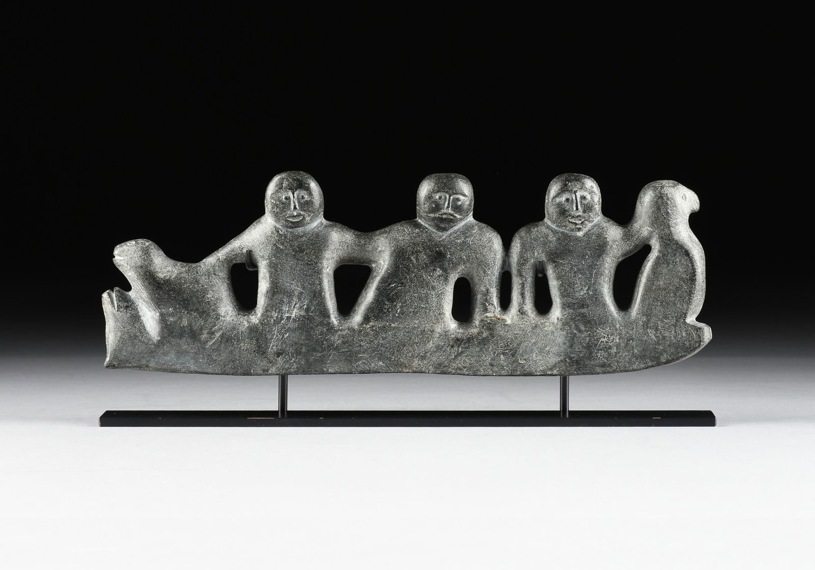 OTTOCHIE ASHOONA (Inuk 1942-1970) "Figure Group," 1960s,: OTTOCHIE ASHOONA (Inuk 1942-1970) "Figure Group," 1960s,a carved stone sculpture of three figures flanked by animals, together with a custom metal stand.5 1/2 x 16 1/2 x 1 1/2 in. (14 x 41.9 x 3.8 cm.