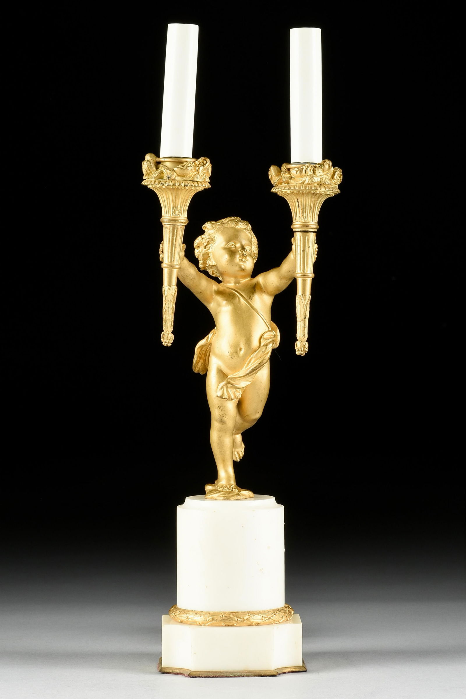 AN NEOCLASSICAL STYLE BRONZE ORMOLU AND MARBLE PUTTO LAMP, FRENCH, 20TH CENTURY,: AN NEOCLASSICAL STYLE BRONZE ORMOLU AND MARBLE PUTTO LAMP, FRENCH, 20TH CENTURY,a gilt bronze putto holding two torch lights mounted on a white marble base.21 1/4 x 5 3/4 x 5 in. (54 x 14.6 x 12.7 cm.