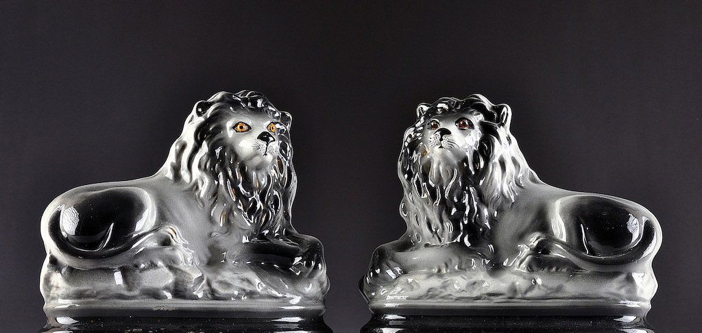 A PAIR OF STAFFORDSHIRE BLACK AND GRAY GLAZED EARTHENWA: A PAIR OF STAFFORDSHIRE BLACK AND GRAY GLAZED EARTHENWARE LION FIGURES, 19TH CENTURY, each modeled in a recumbent position with glass eyes and resting on an incorporated oval molded plinth, parcel gil