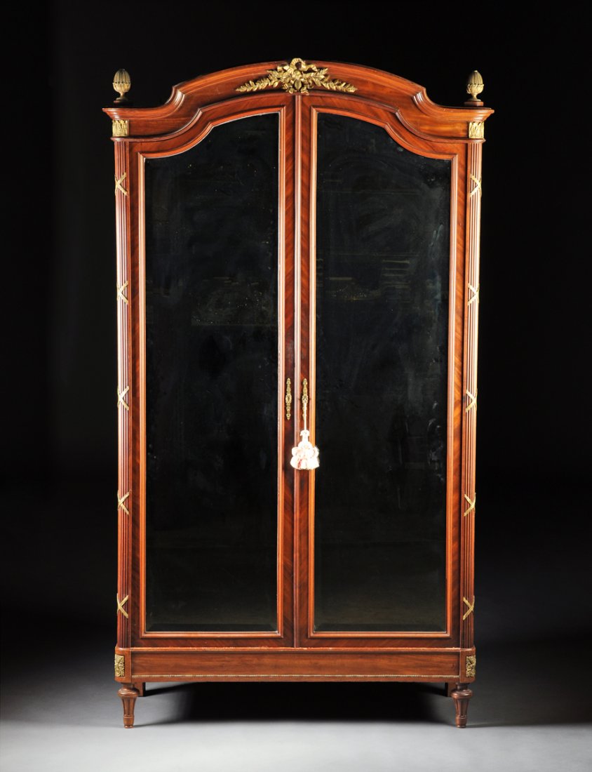 A LOUIS XVI STYLE GILT BRONZE MOUNTED MAHOGANY ARMOIRE: A LOUIS XVI STYLE GILT BRONZE MOUNTED MAHOGANY ARMOIRE WITH MIRROR PANELED DOORS, LATE 19TH/EARLY 20TH CENTURY, with an arched molded cornice centering a ribbon tied berried laurel branch mount, flank