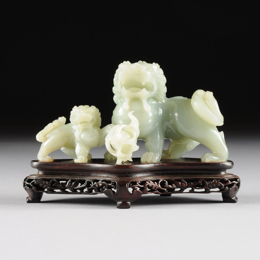 A CHINESE CARVED JADEITE FIGURAL GROUP OF TWO FOO DOGS,: A CHINESE CARVED JADEITE FIGURAL GROUP OF TWO FOO DOGS, 20TH CENTURY, of a large foo dog with a small one to his side, joined by a puzzle ball, of gray/green jadeite, mounted on carved wooden base. He