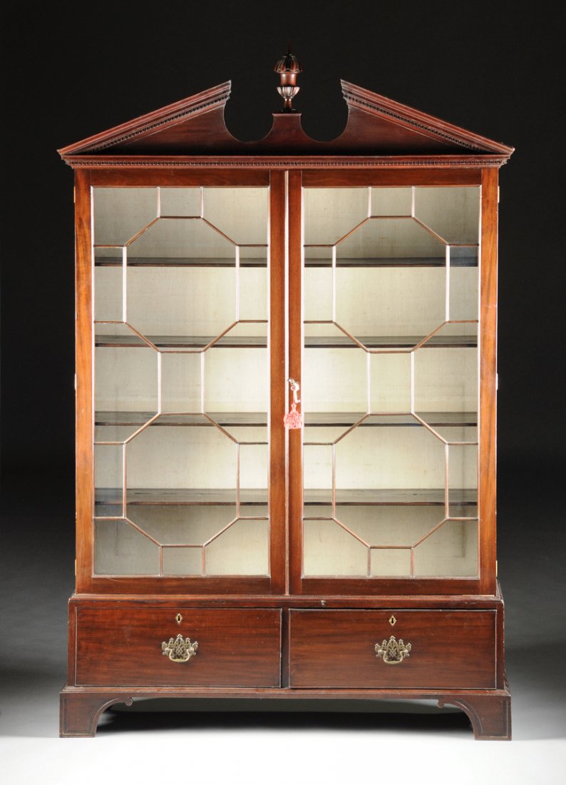 A GEORGE III CARVED MAHOGANY BOOKCASE CABINET, CIRCA: A GEORGE III CARVED MAHOGANY BOOKCASE CABINET, CIRCA 1800, with a dentilated broken pediment cornice centering a gadrooned urn finial, above a dentilated frieze and paired glazed doors enclosing octag