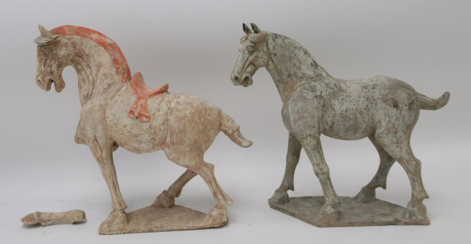 Two Large Tang Style Horses: The large saddled horse, well molded with white and red pigment has one foreleg raised (broken but retained). Offered with a smaller horse of white/grey pigment. Both showing some repair. 18" and 17" 