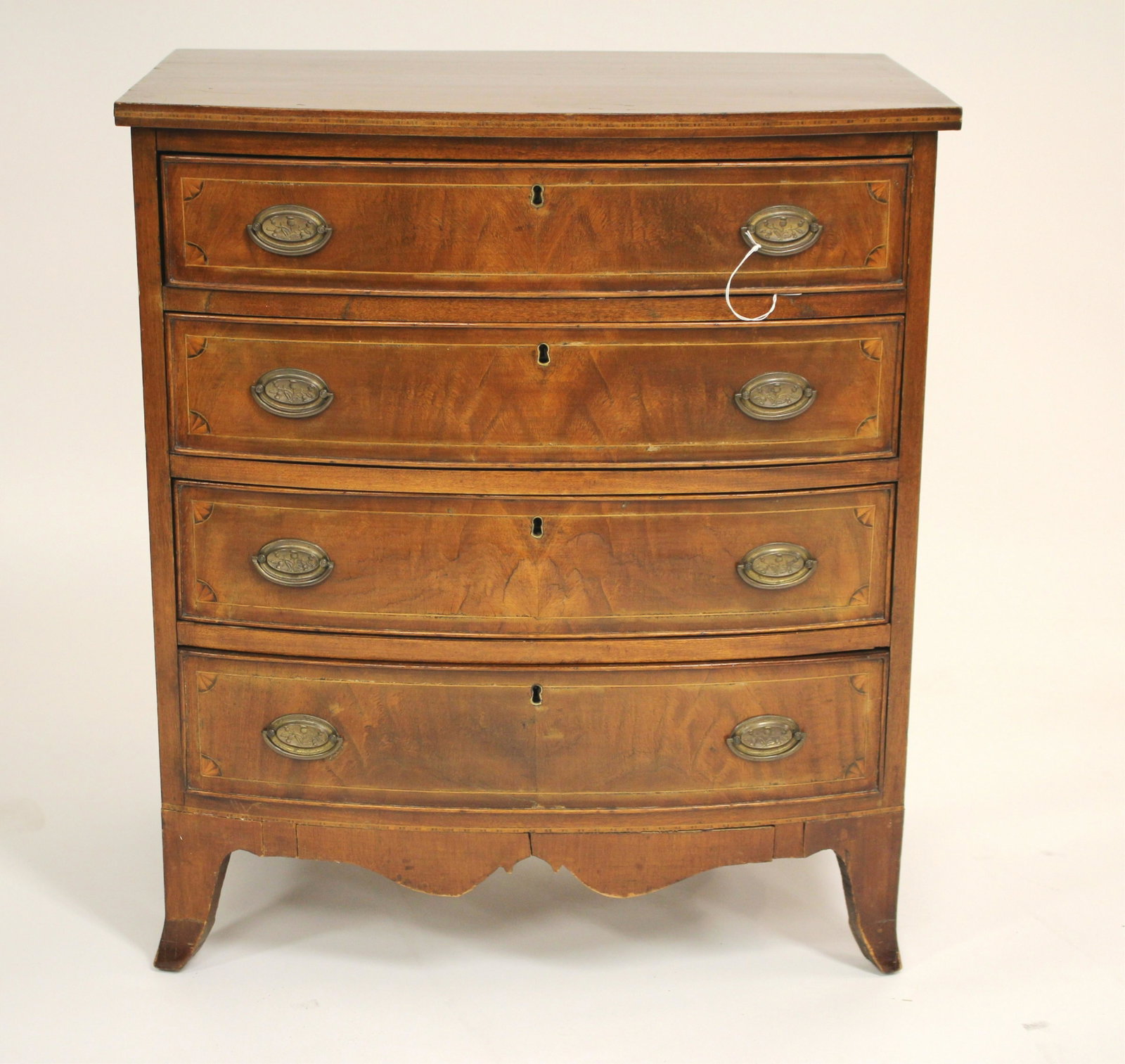 Diminuitive Hepplewhite Bowfront Chest of Drawers: Veneer details include drawer face corner fans; pressed brass thistle decoration bail handles; lroughly, freehand carved base apron, possibly done later and /or as part of an alteration 27.5"W x 18.25