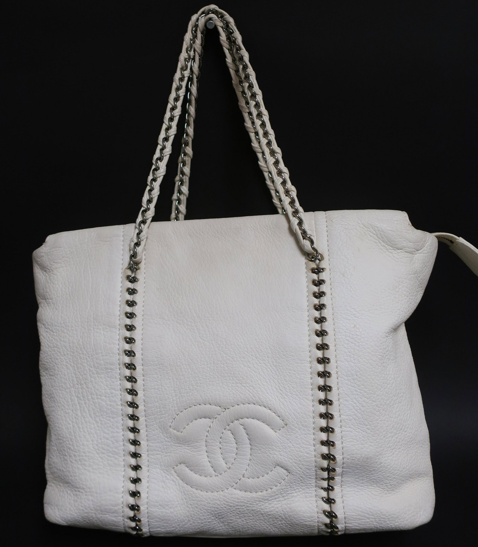 Chanel Grained Leather Lambskin Tote: White soft grained leather exterior with white lambskin smooth leather interior. Two slip pockets, one zip pocket. Zip top closure. Silver hardware. Braided chain handles with same detail on front and
