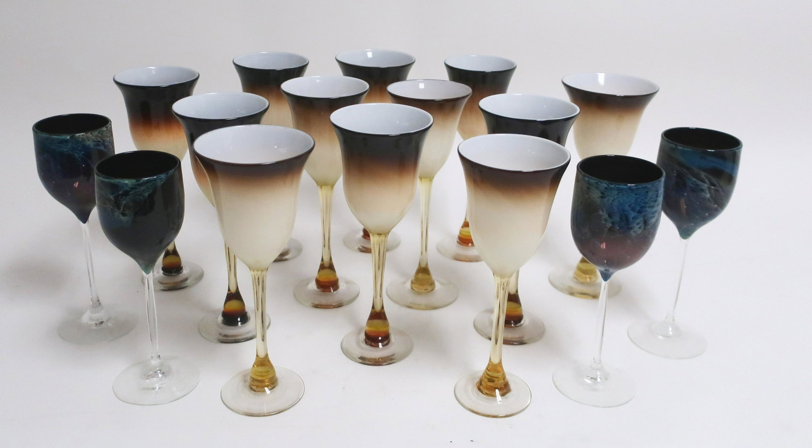16 Blown Art Glass Stemmed Wine Glasses: (4) in celestial "windswept" blue pattern, signed Simpson. (12) in opaque mocha/cream fade, espresso rim, white goblet interior; some variations in goblet size. 8.5" & 9.5H (QDRIC9452/16)(JP)