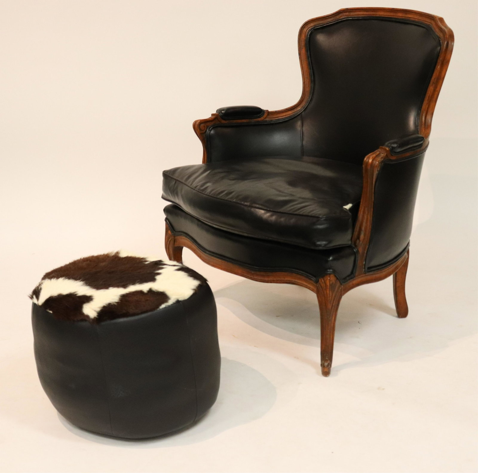 Black Leather Bergere and a Pouf: Carved wood frame with leather upholstery, down and feather cushion. Antiqued stain finish with faux worm holes, wood dents. Leather pouf with hide top. (WECH2812/2)(JP) Dimensions: 35"H x 28.5W x 21 