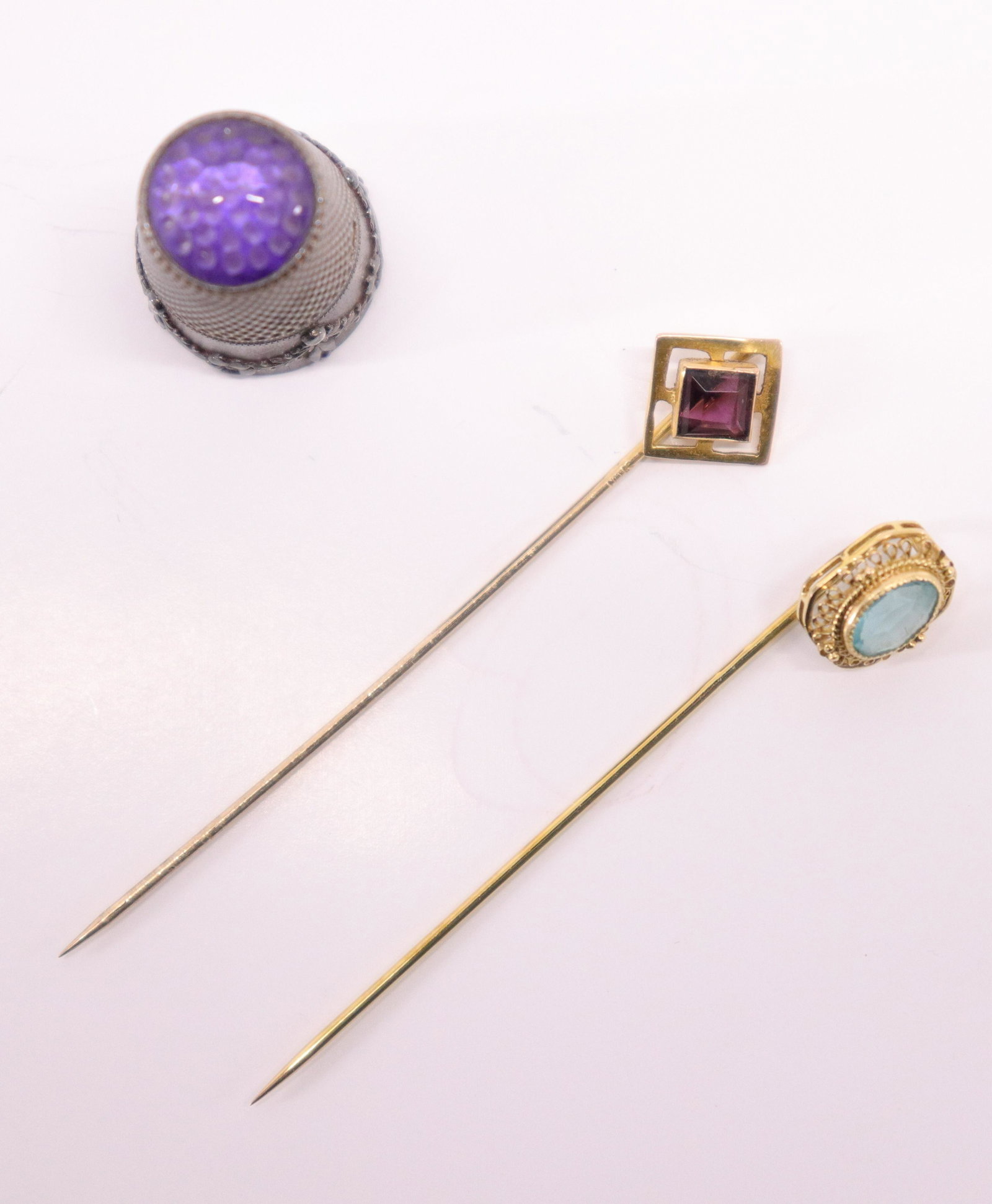 800 Silver Amethyst Mounted Thimble, 2 Stickpins: Thimble lower body with floral swags. Punctuated amethyst stone top. Side marked 800. 10K gold stickpins pins, one aquamarine and one garnet color glass stones. Dimensions: Thimble 1"H Stickpins 2.25 