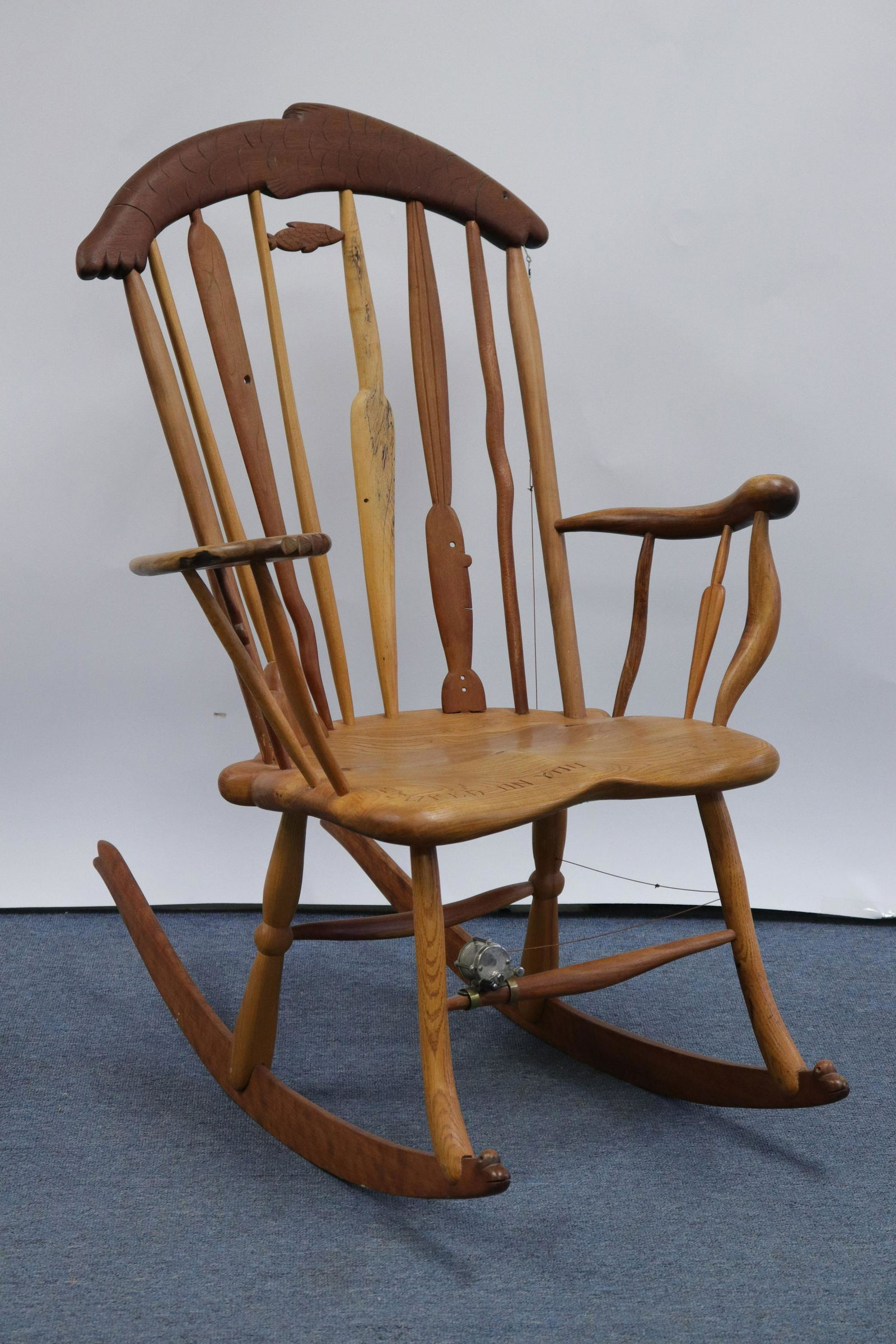 Tommy Simpson "Hooked On You" Fishing Theme Rocker: L20C. Mixed woods. Asymmetrical arms. Metal fishing reel with line. Wood inlays. Heart shaped wood-check butterfly. Fish form crest rail. "Hooked On You" inscribe carved into seat. Signed Tommy S unde