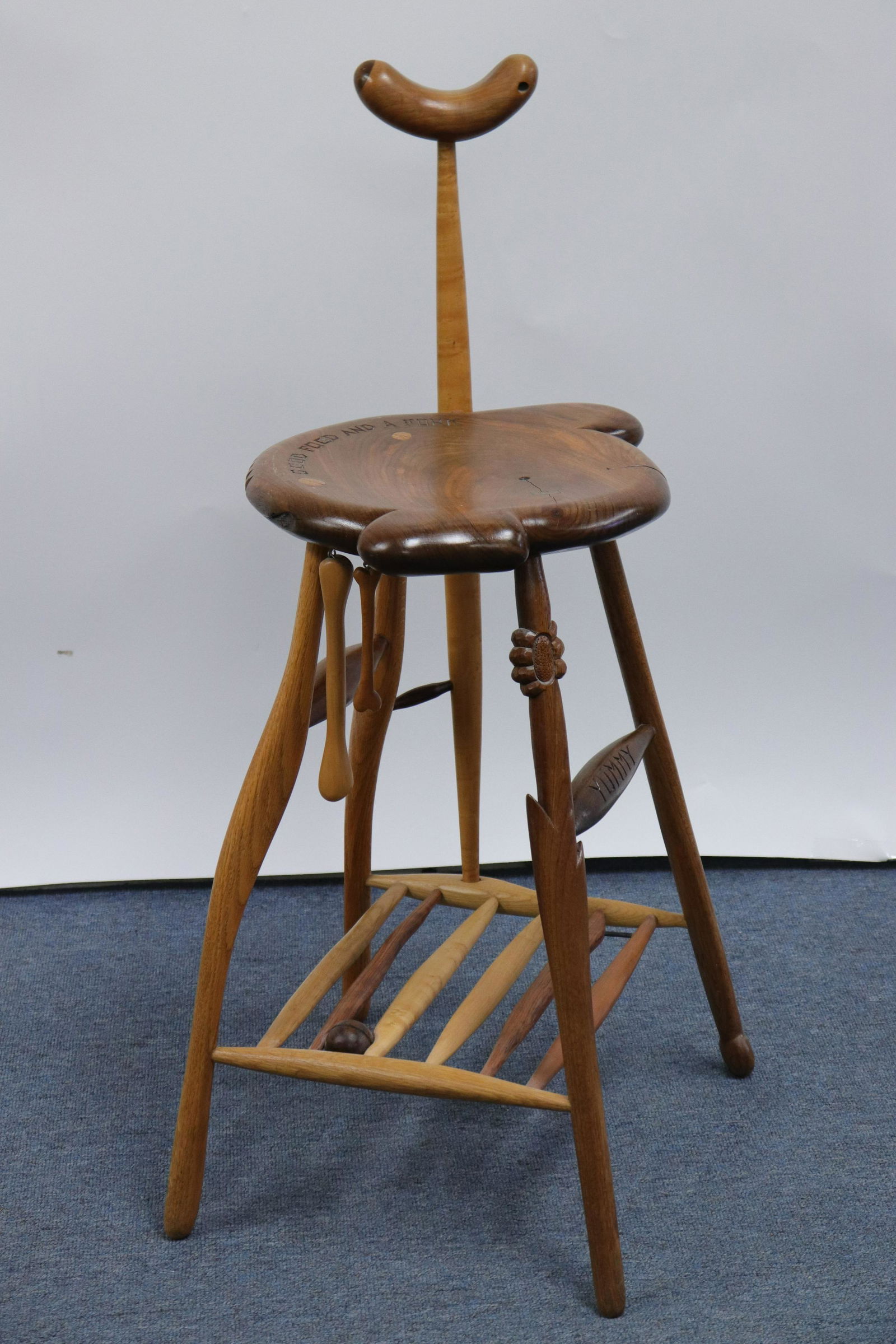 Tommy Simpson "Good Food And A Fork" Stool: L20C. As kitchen/bar/task seating. Mixed woods. Yoke on turned spindle with tenon and key joint armrest. Shaped seat with carved notches,inscribe carved "Good Food And A Fork" and "Yummy". Two danglin