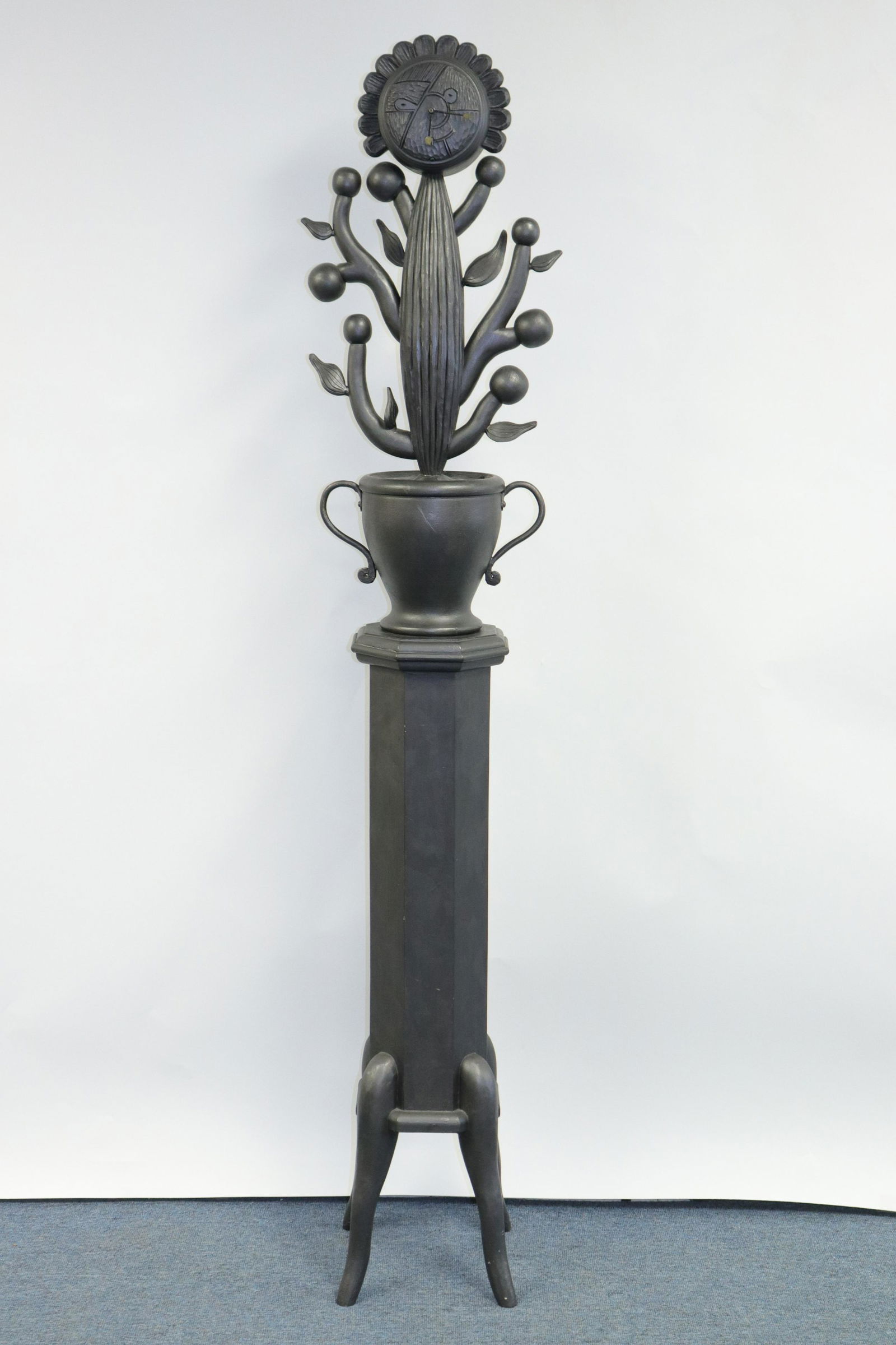 Tommy Simpson Flower Clock Sculpture: L20C. Carved wood. Two sections: flowerpot with flower face clock top on four legged octagonal pedestal. Painted in grey-black/silvery grey-black. Removable back with battery operated clock. Dimension