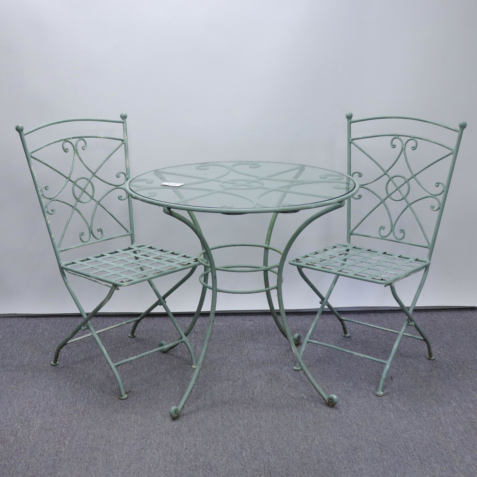 Vintage Painted Metal Bistro Table, Folding Chairs: Splayed leg base with glass top insert. Two matching design side chairs. Scrolled and cross bar patterns. Grey-green paint. Strong, heavy material. Dimensions: 29.75"H x 31.25 dia. chair 17H @ seat x