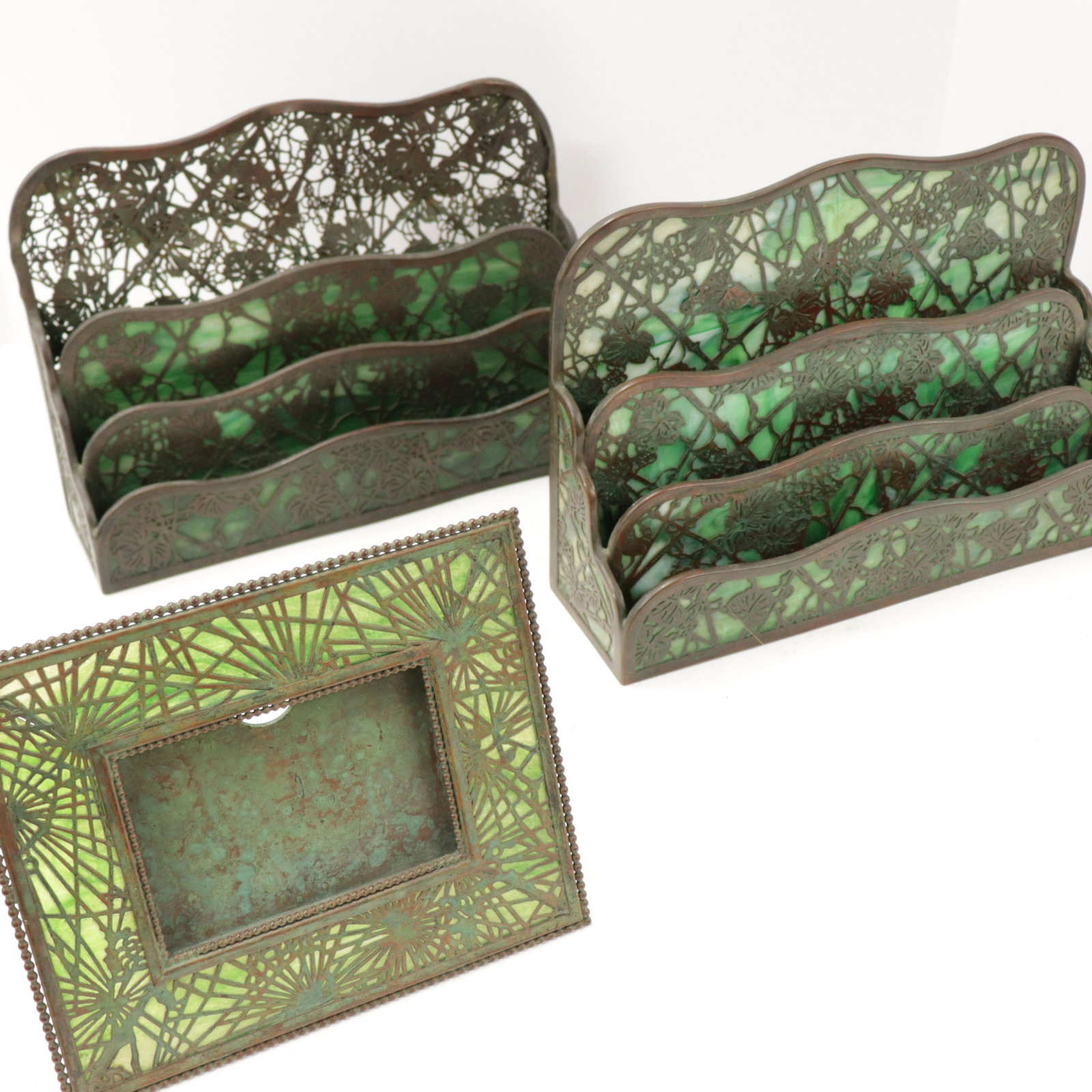 3 Tiffany Studios Bronze & Favrile Desk Items: 3 Tiffany Studios Bronze & Favrile Glass Desk Articles, comprises: 2 Grapevine pattern paper racks and a Pine Needle pattern calendar frame Dimensions: 8 1/2" x 12 1/2" and 7 1/" x 8 3/4" Property fro