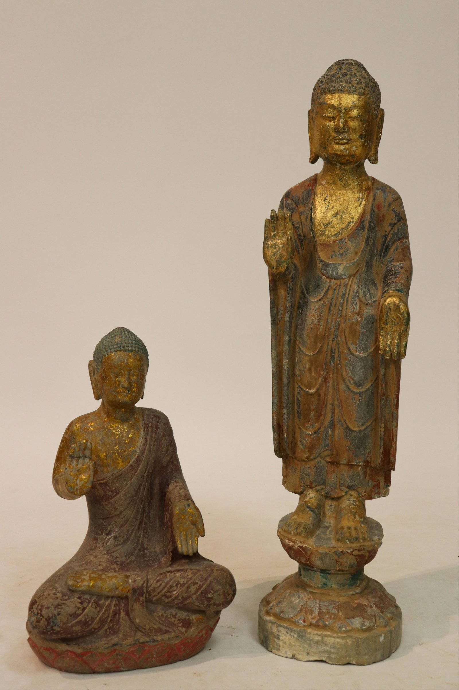 Two Stone Buddha Statues: One standing and one sitting, both are solidly on lotus plinths and in the combination of abyhaya and varada mudra. Each has a pleasing patina of paint and gilt applied to the surfaceNOTE: This lot is