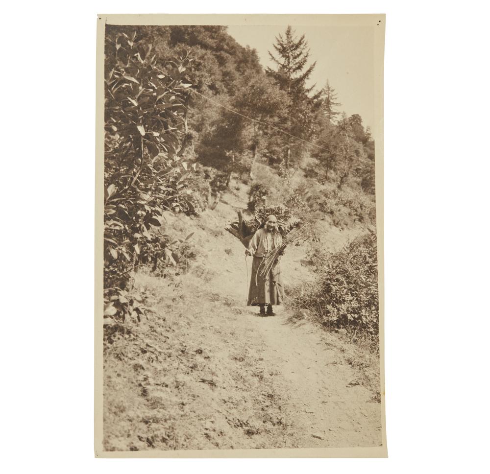 Grace Nicholson Photograph of Karok Indian: Photograph of 90 year old Karok woman carrying a load of wood by Grace Nicholson. Handwritten in ink description verso. Stamped, verso, "Photo by Grace Nicholson, Pasadena, Cal." Dimensions: (overall)