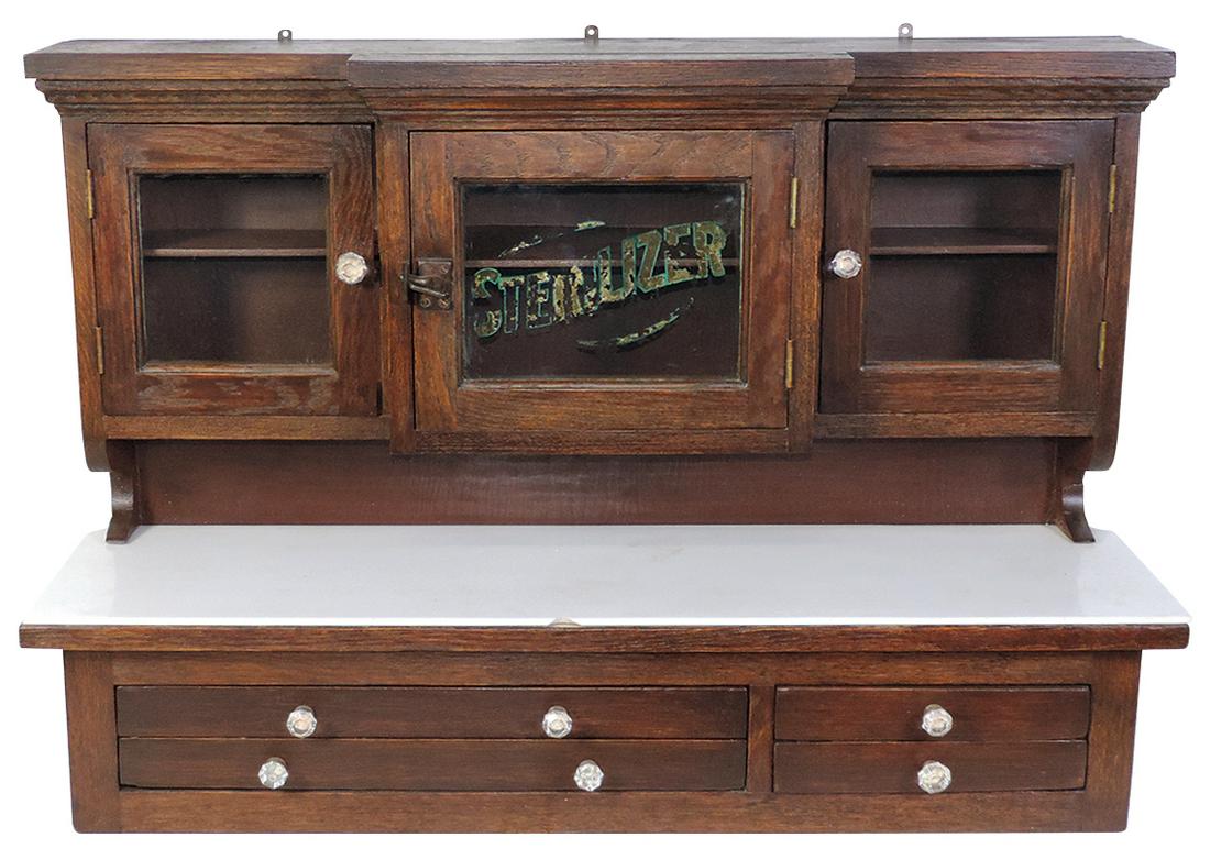 Barber Shop Wall Cabinet, oak w/3 set-back glass doors w/gilt "Sterilizer" over milk glass top w/4: Barber Shop Wall Cabinet, oak w/3 set-back glass doors w/gilt "Sterilizer" over milk glass top w/4 shallow drawers, chip to glass top, o/wise VG+ customization, 24.75"H x 36"W x 10"D.