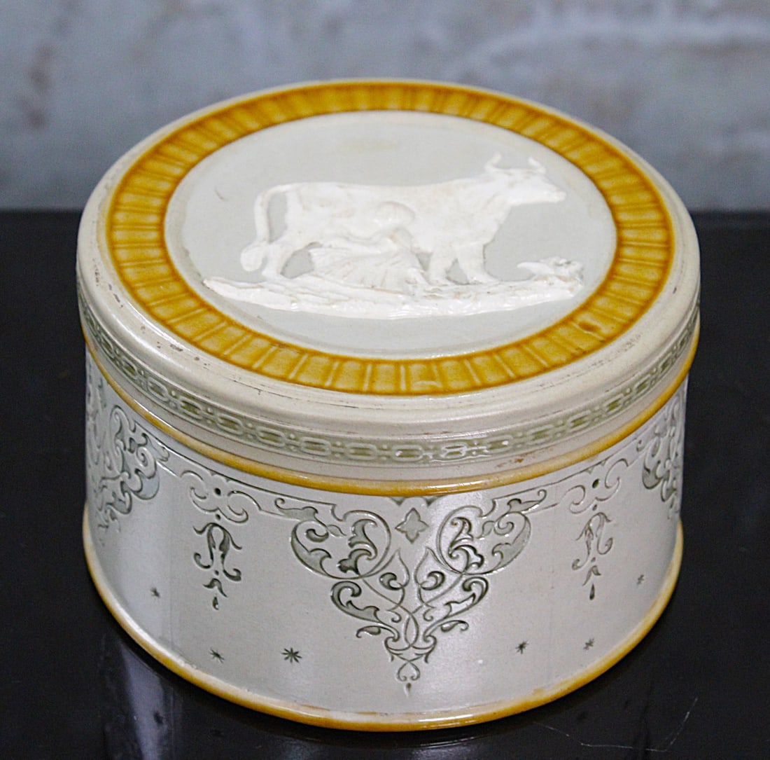 A German Mettlach Stoneware Box: Embossed scene of a maiden milking a cow on cover, Mettlach backstamp and incised 2115, VG+ cond with small flake on rim, 4.75"Dia.
