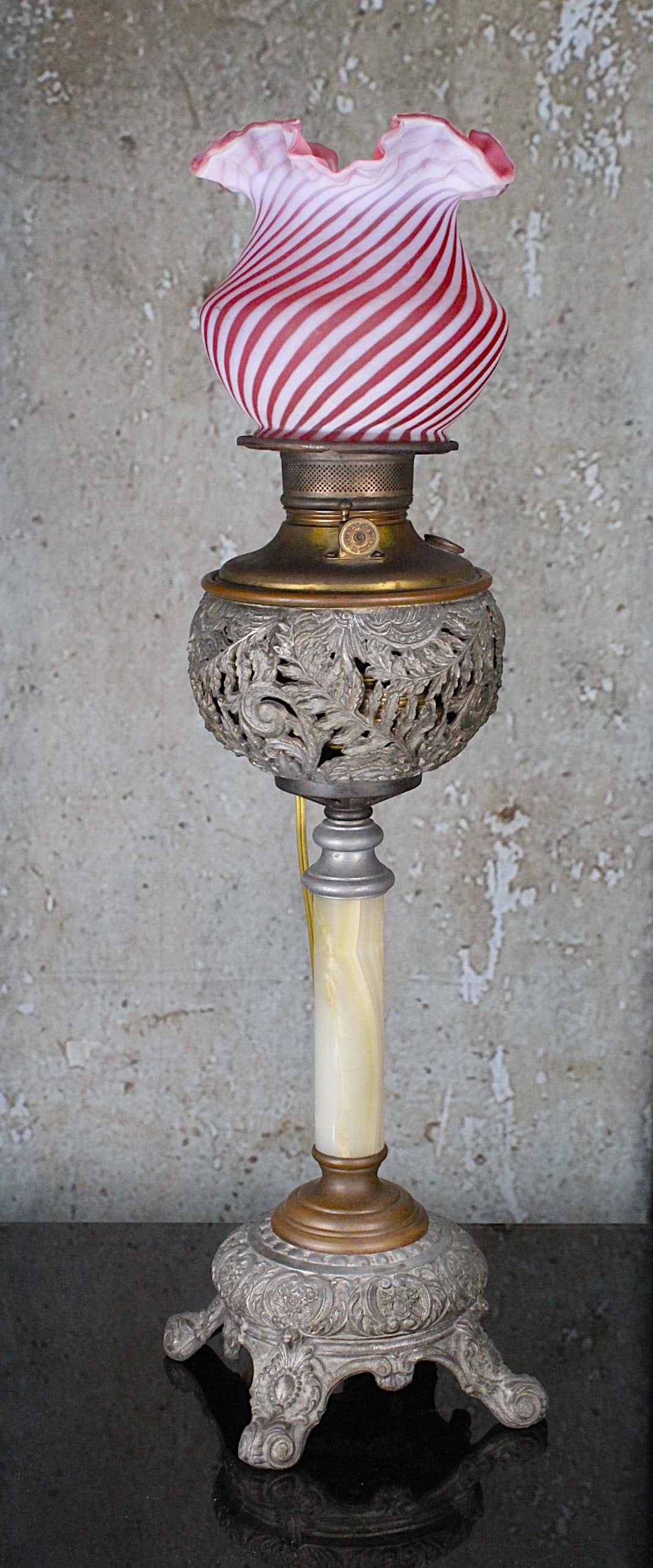 A Victorian Filigree Banquet Lamp: Onyx stem on embossed metal plinth supporting a filigree font with associated opalescent cranberry swirl shade, likely by Edward Miller, marked ?Made in U.S. Of America? G+ working cond w/later elec. 